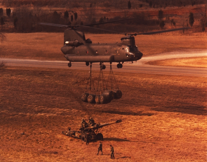 A photograph of an early CH-47D transporting fuel blivets near an M198 Howitzer.